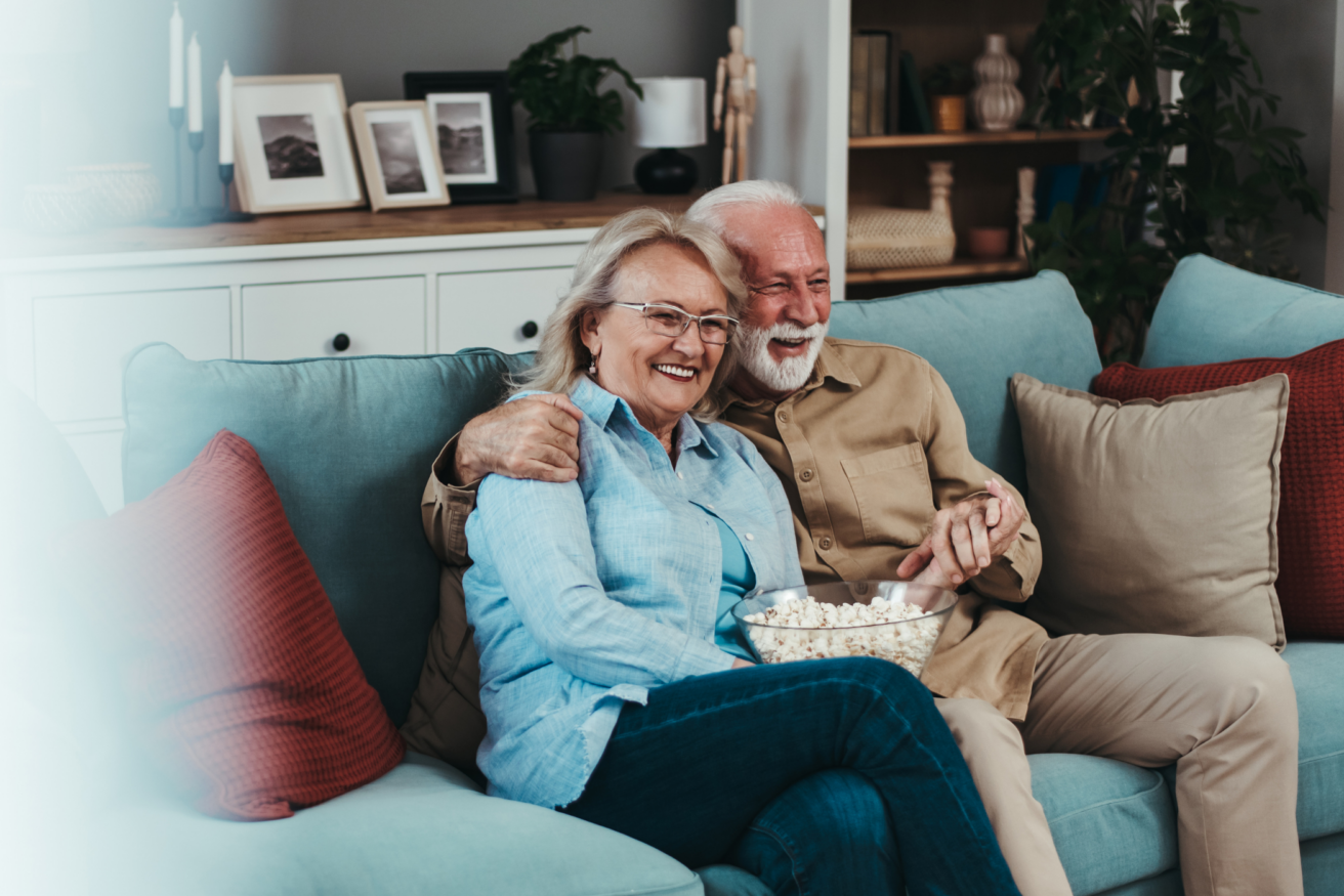 Elderly couple sitting on a couch, smiling while sharing a bowl of popcorn in a cozy living room setting with shelves and framed pictures in the background.