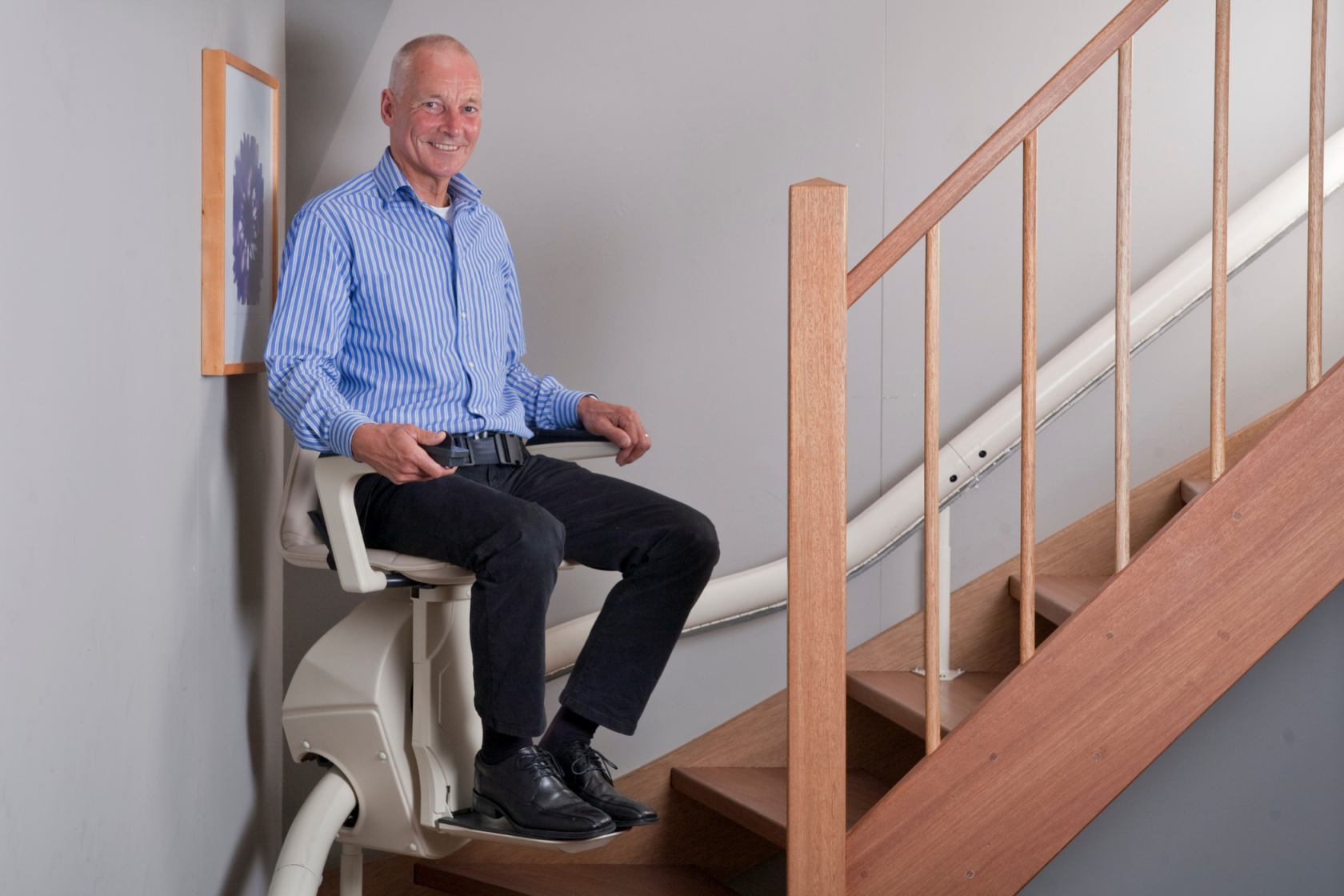 A man in a blue striped shirt and black pants uses a stairlift to ascend a wooden staircase inside a home.