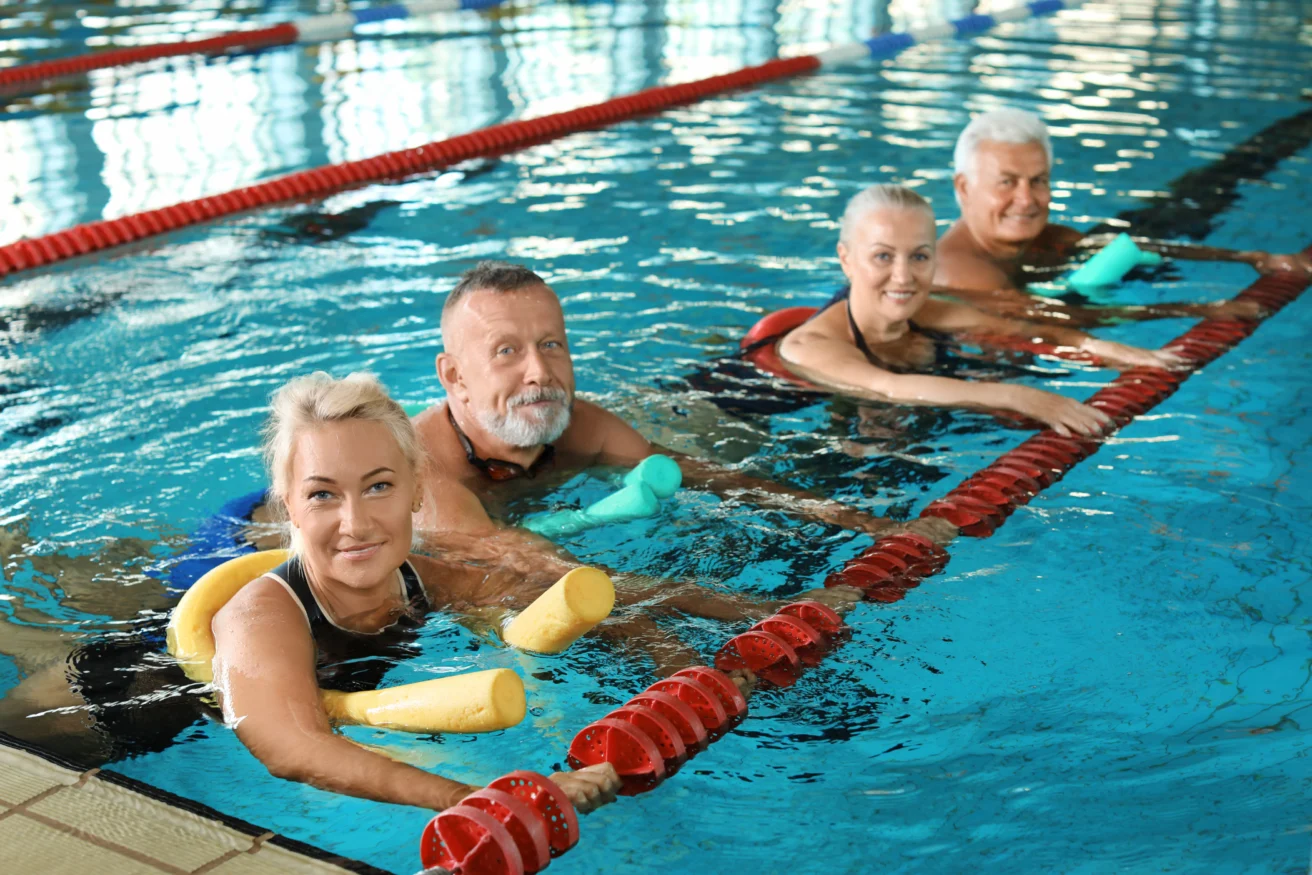 Four smiling older adults in a swimming pool, using foam noodles for support, hold onto the lane divider.