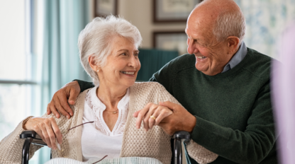 An elderly woman in a wheelchair and an elderly man are smiling and holding hands, sitting close in a well-lit room. The woman holds her glasses in her other hand.