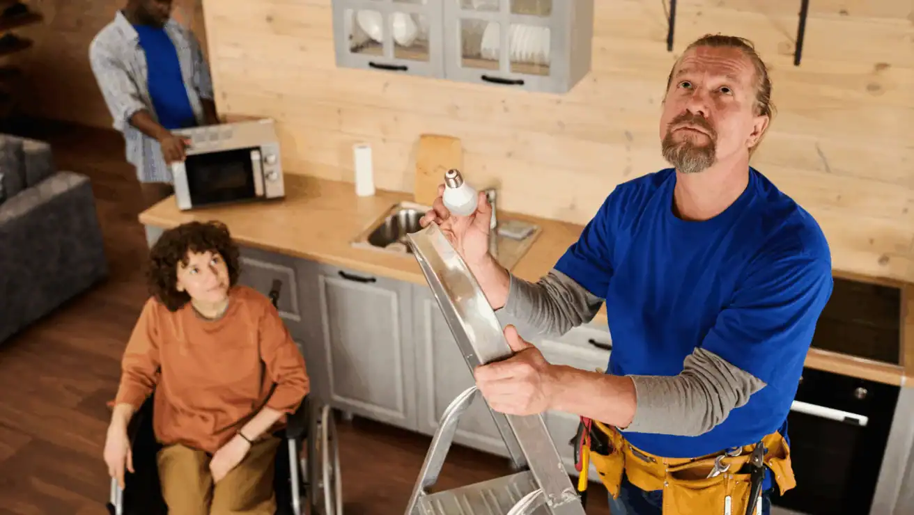A man on a ladder replaces a lightbulb in a kitchen while a woman in a wheelchair and another man by the counter look on.