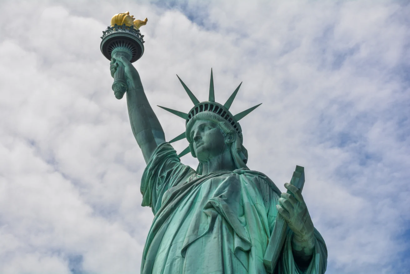 A close-up view of the Statue of Liberty against a cloudy sky, showing the torch in her raised right hand and a tablet in her left.