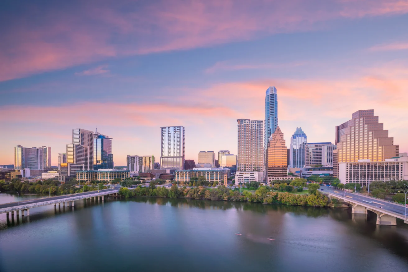 A city skyline at sunrise featuring tall buildings and a calm river in the foreground with a bridge crossing it.