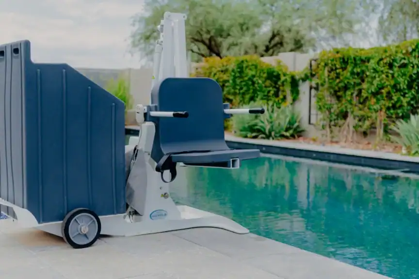A pool lift chair is positioned at the edge of an outdoor swimming pool, with greenery and a fence in the background.