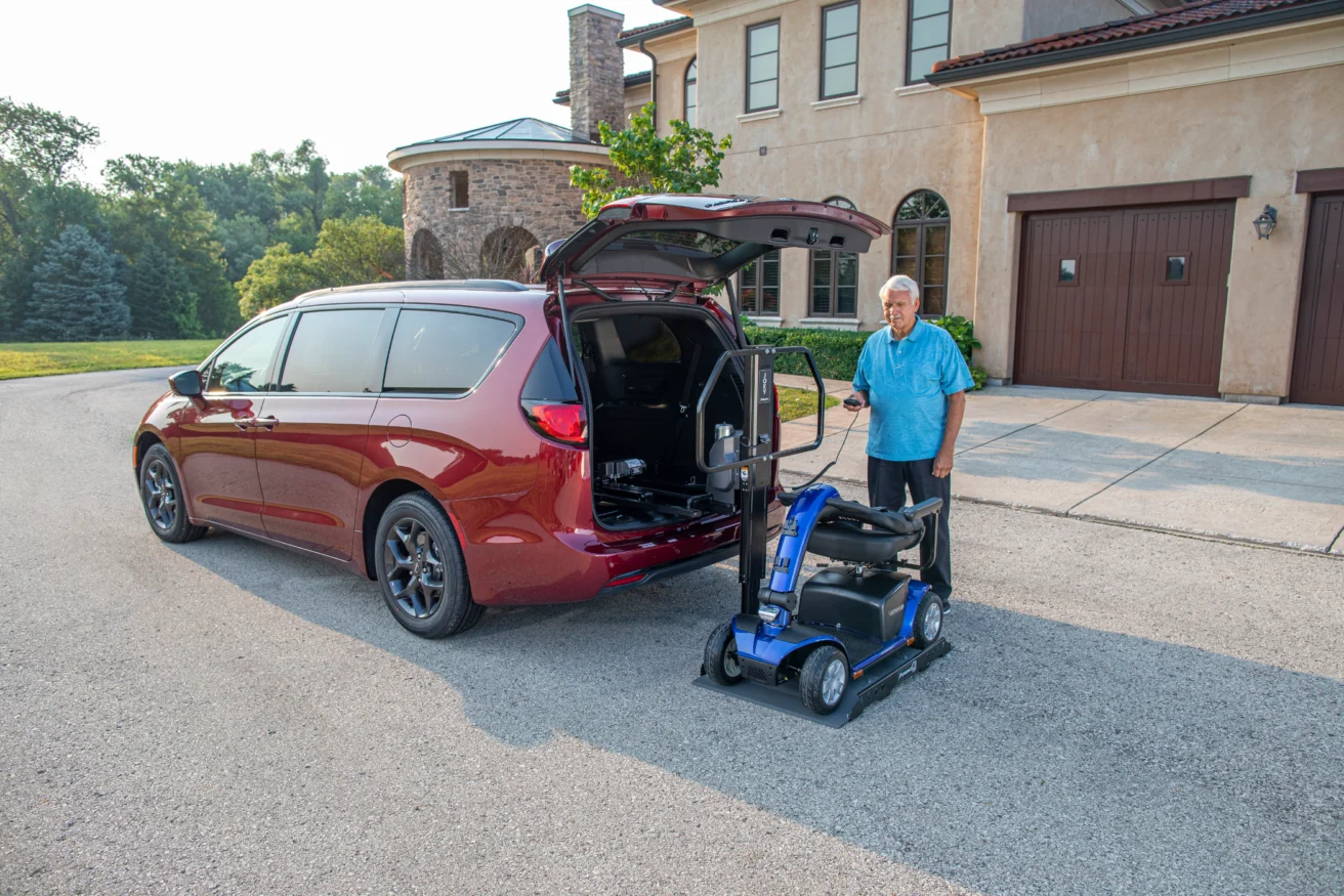An elderly man standing next to an open minivan, assisted by a mobility scooter lift positioned at the rear of the vehicle.