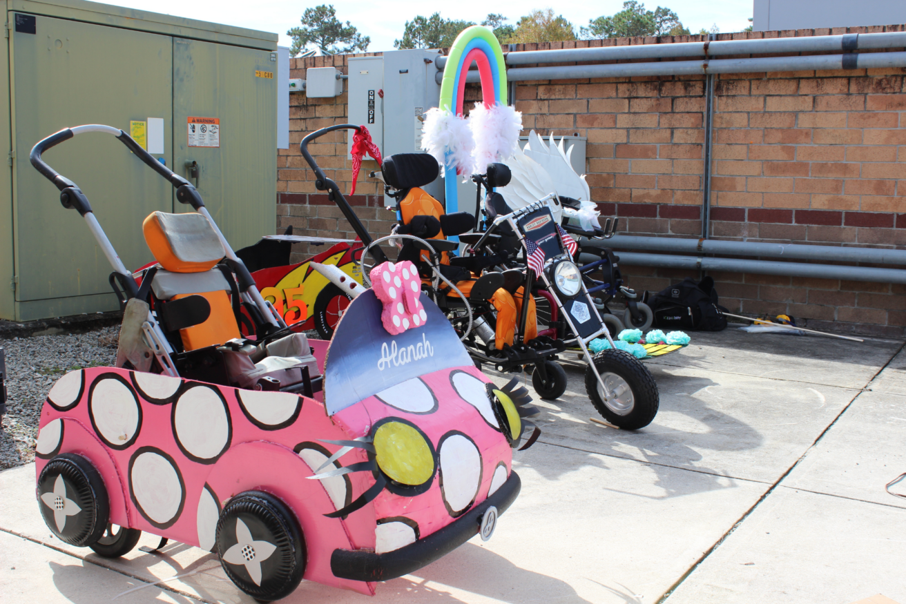 Three decorated children's wheelchairs are parked outside a building: one pink with polka dots, another black with an American flag, and a third with a rainbow and white wings.