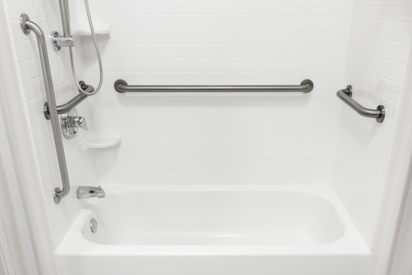 A white bathroom featuring a bathtub, shower fixtures, horizontal grab bars on three sides, and a small corner shelf.