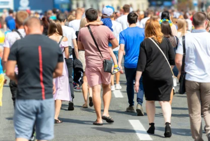 A crowd of people, including a man carrying a child, walk on a sunny street. The group consists of individuals wearing various casual summer clothes.