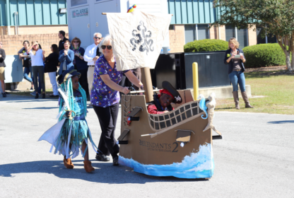 A woman pushes a child in a pirate-themed wheelchair while another child dressed in a costume walks alongside. Several people watch in the background.