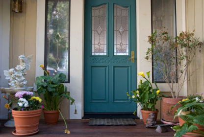 A teal door with decorative glass panels is flanked by two tall windows. Potted plants and flowers are arranged on the wooden porch, along with a frog statue and a white angel sculpture.