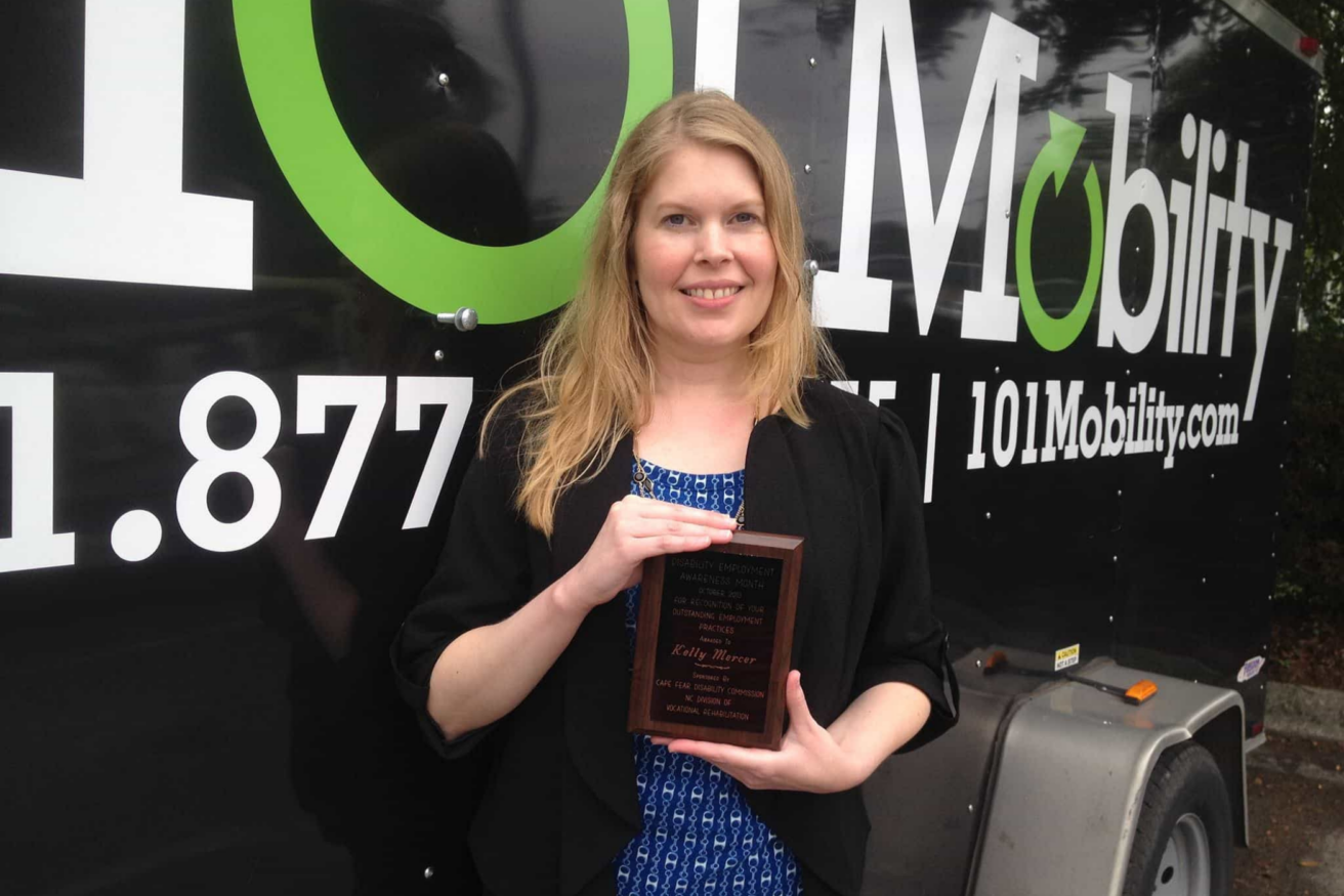 A woman stands in front of a 101 Mobility trailer, holding a plaque.