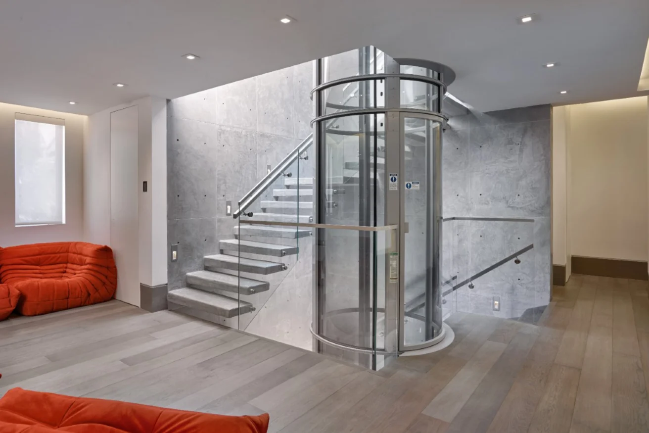 Modern interior featuring a spiral glass elevator and a sleek staircase with metal railings, surrounded by light gray walls and wood flooring, with red seating in the corner.