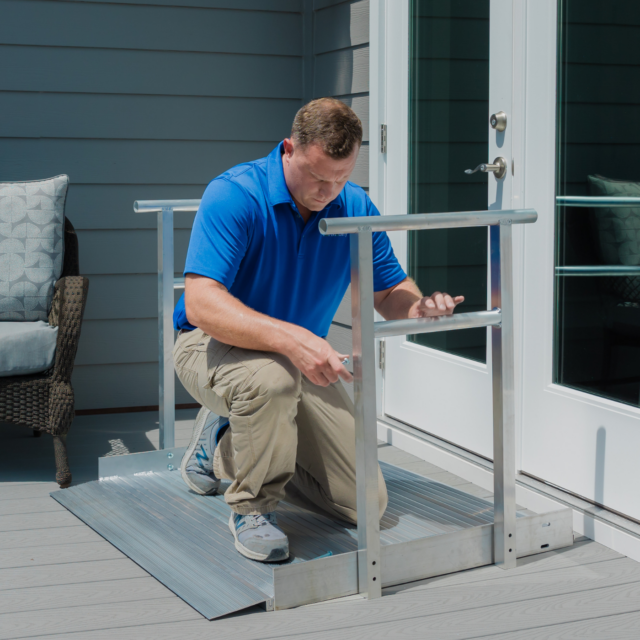 A man in a blue shirt installs a metal wheelchair ramp at the entryway of a home. A wicker chair and a cordless drill are nearby.