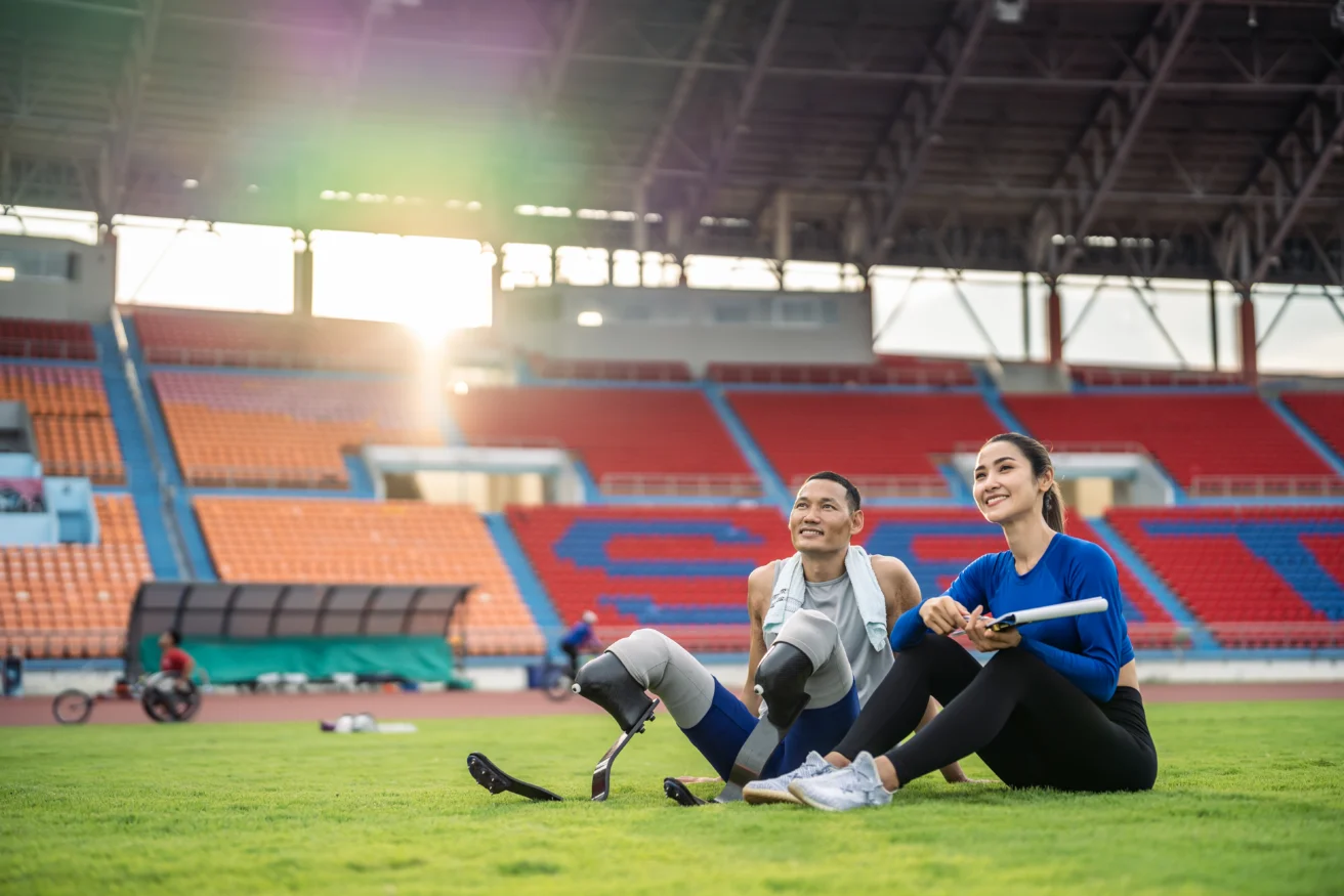Two athletes with prosthetic legs sit on grass in a stadium, smiling and looking ahead. Sunlight filters through the stands.