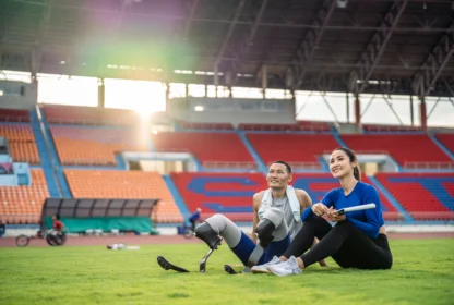 Two athletes with prosthetic legs sit on grass in a stadium, smiling and looking ahead. Sunlight filters through the stands.