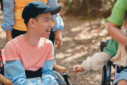 A person in a wheelchair wearing a pink shirt and blue cap smiles outdoors, surrounded by others.