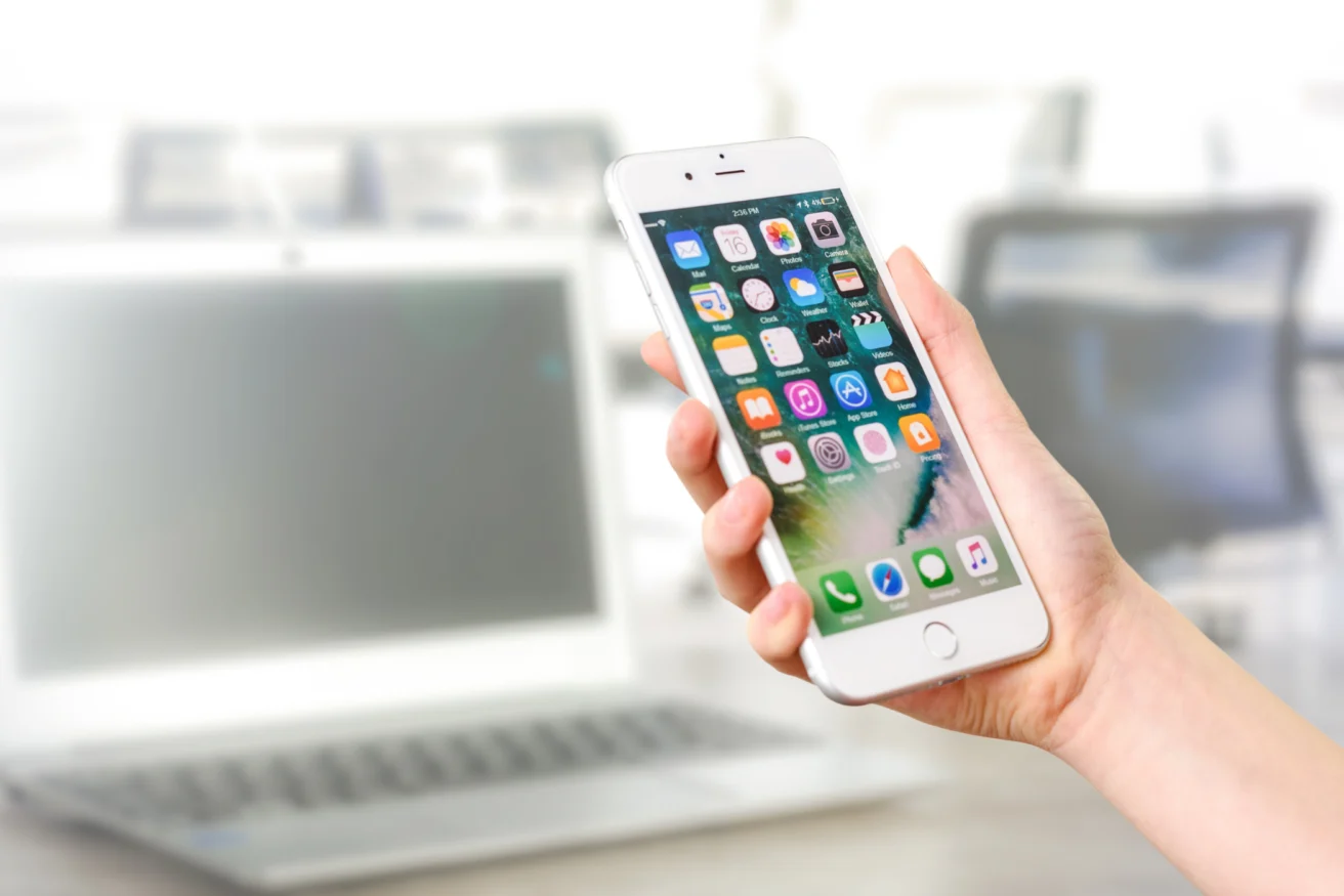 A hand is holding a smartphone displaying several app icons, with a blurred laptop in the background.