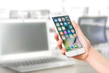 A hand is holding a smartphone displaying several app icons, with a blurred laptop in the background.