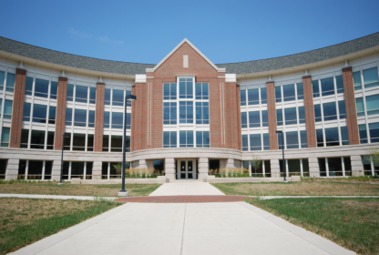 A modern brick and glass building with a curved facade and a central entrance, surrounded by grass and a paved walkway under a clear blue sky.