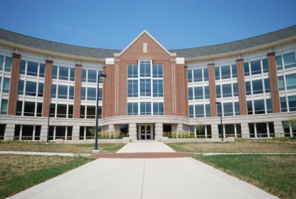 A modern brick and glass building with a curved facade and a central entrance, surrounded by grass and a paved walkway under a clear blue sky.