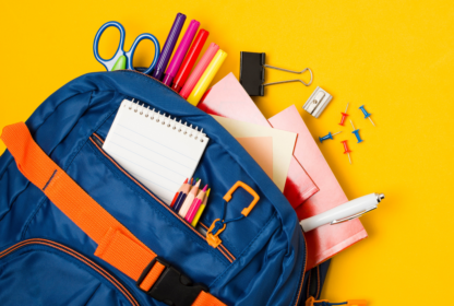 A blue backpack with orange accents contains colorful pens, a notepad, scissors, papers, a pen, a binder clip, a sharpener, and push pins, all set against a yellow background.