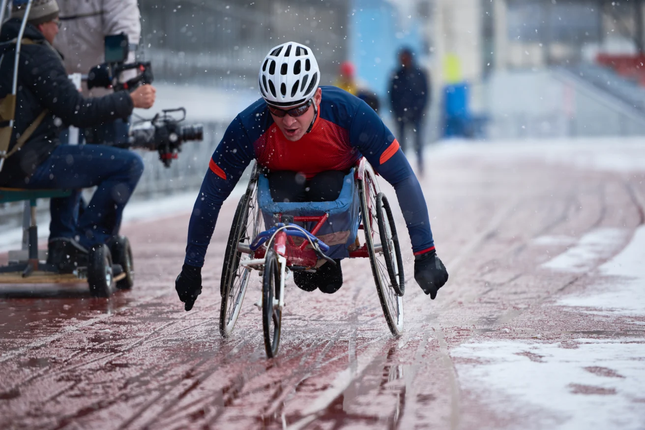 A person in a racing wheelchair competes on a snowy track, wearing a helmet and winter athletic gear. A cameraman is filming to the side.