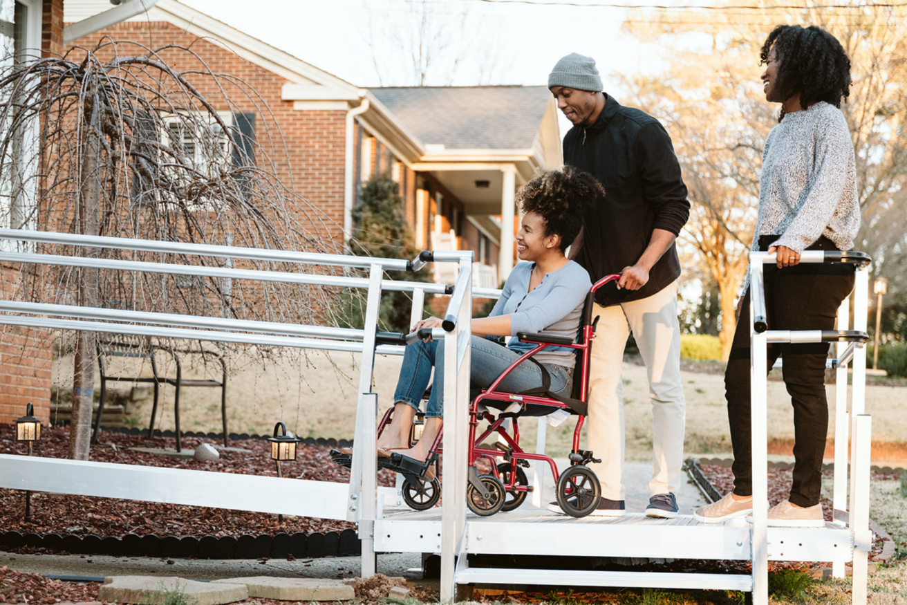 A person in a wheelchair is being pushed up a ramp by another individual. Another person stands beside them, smiling. A brick house is in the background.