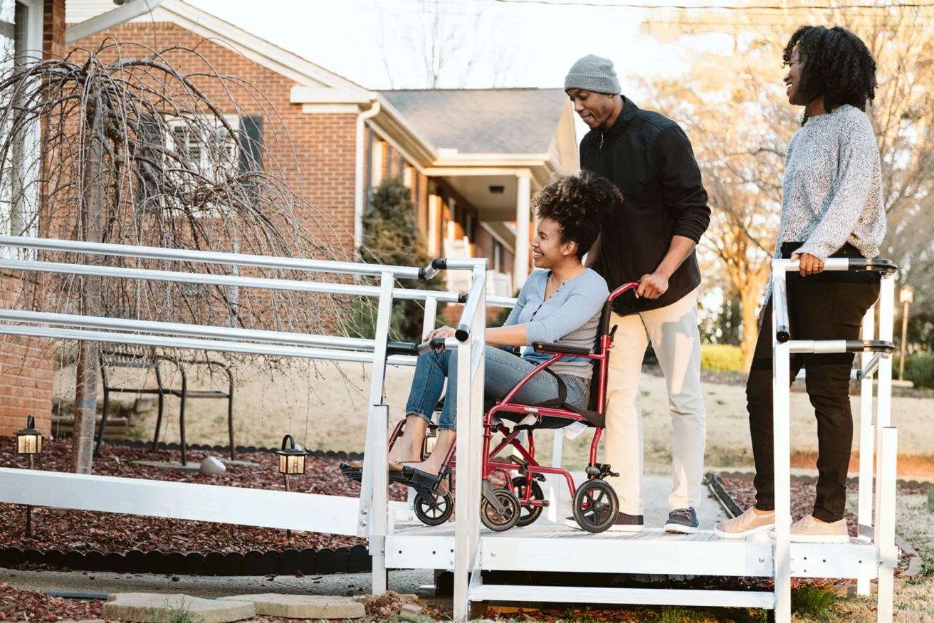 A person in a wheelchair is being pushed up a ramp by another individual. Another person stands beside them, smiling. A brick house is in the background.