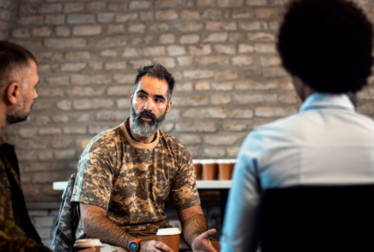 A man in a camouflage shirt is talking to two individuals in front of a brick wall. He holds a paper cup while engaged in conversation.