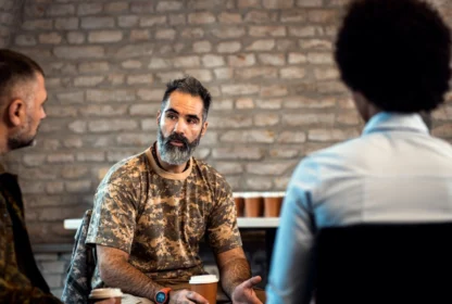 A man in a camouflage shirt is talking to two individuals in front of a brick wall. He holds a paper cup while engaged in conversation.