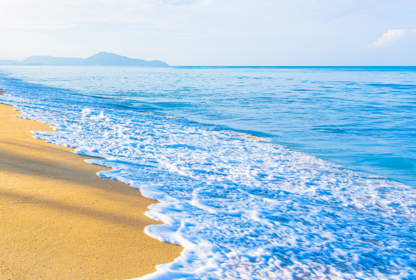 A calm beach scene with gentle waves washing onto a sandy shore, under a clear sky with distant mountains in the background.