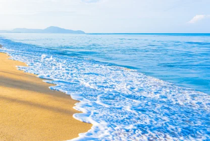 A calm beach scene with gentle waves washing onto a sandy shore, under a clear sky with distant mountains in the background.