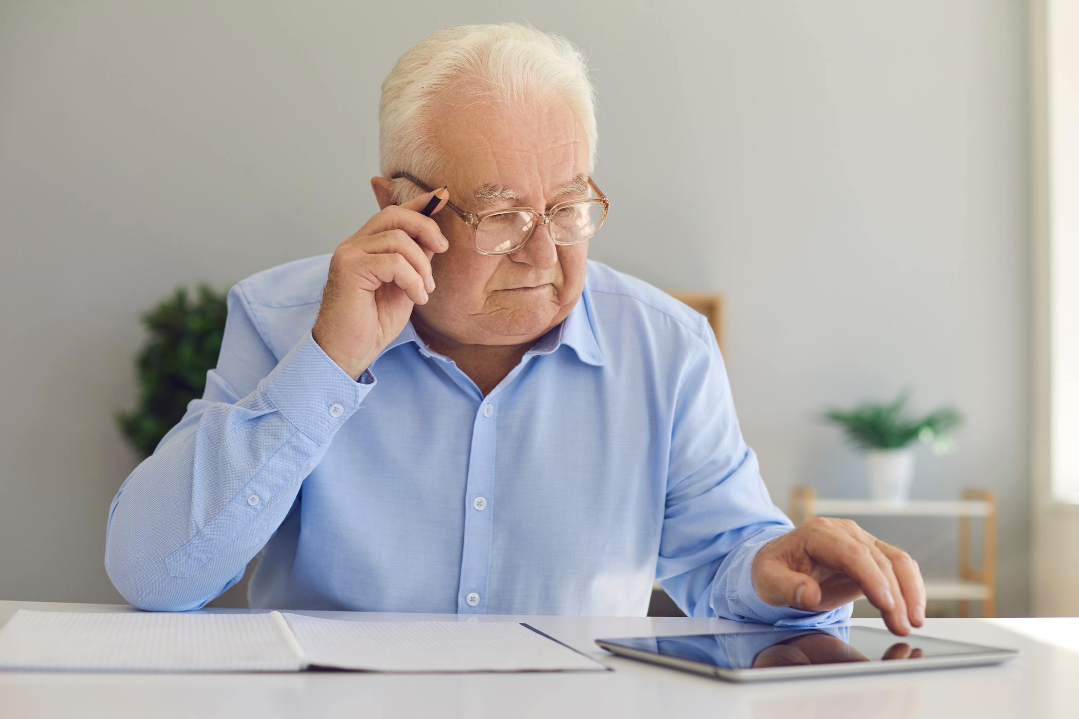 Elderly man with glasses intently using a tablet while holding a pen. He is seated at a desk with papers in front of him.