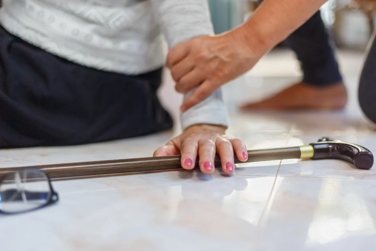 A person helps another who has fallen, with one hand on their arm and a cane and glasses on the floor nearby.