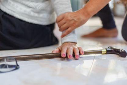 A person helps another who has fallen, with one hand on their arm and a cane and glasses on the floor nearby.