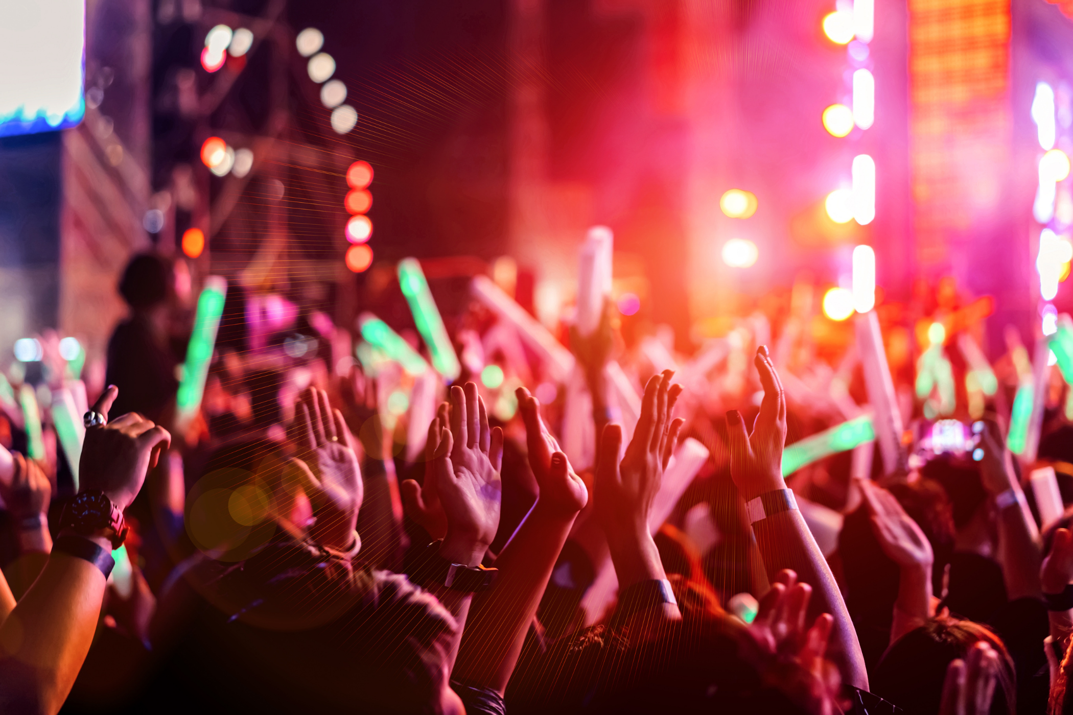 A crowd at a concert raises hands and glow sticks under colorful stage lights, creating a vibrant atmosphere.