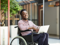 A person in a wheelchair smiles while working on a laptop in a modern indoor setting.