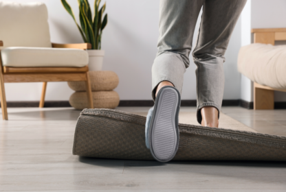 Person tripping over a rolled-up carpet in a modern living room with a wooden chair, a potted plant, and pale wood flooring.