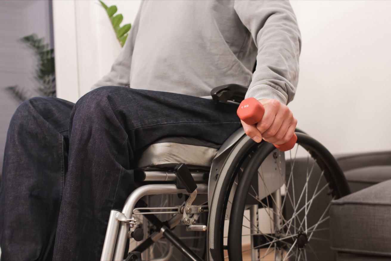 A person in a wheelchair, wearing a grey long-sleeve shirt and jeans, holds a small orange dumbbell.
