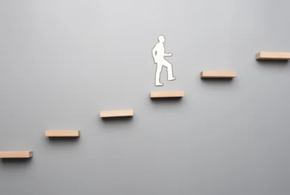 Silhouette of a person ascending a series of floating wooden steps against a plain gray background, symbolizing progress or personal growth.