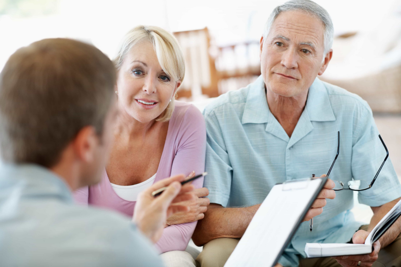 An elderly couple is seated and attentively listening to a man holding paperwork and a pen. The man appears to be explaining or discussing something with them. The couple looks engaged.