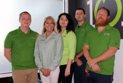 A group of five people, four men and one woman, stand indoors wearing green shirts. They are positioned in front of a wall with the number "101" visible in the background.