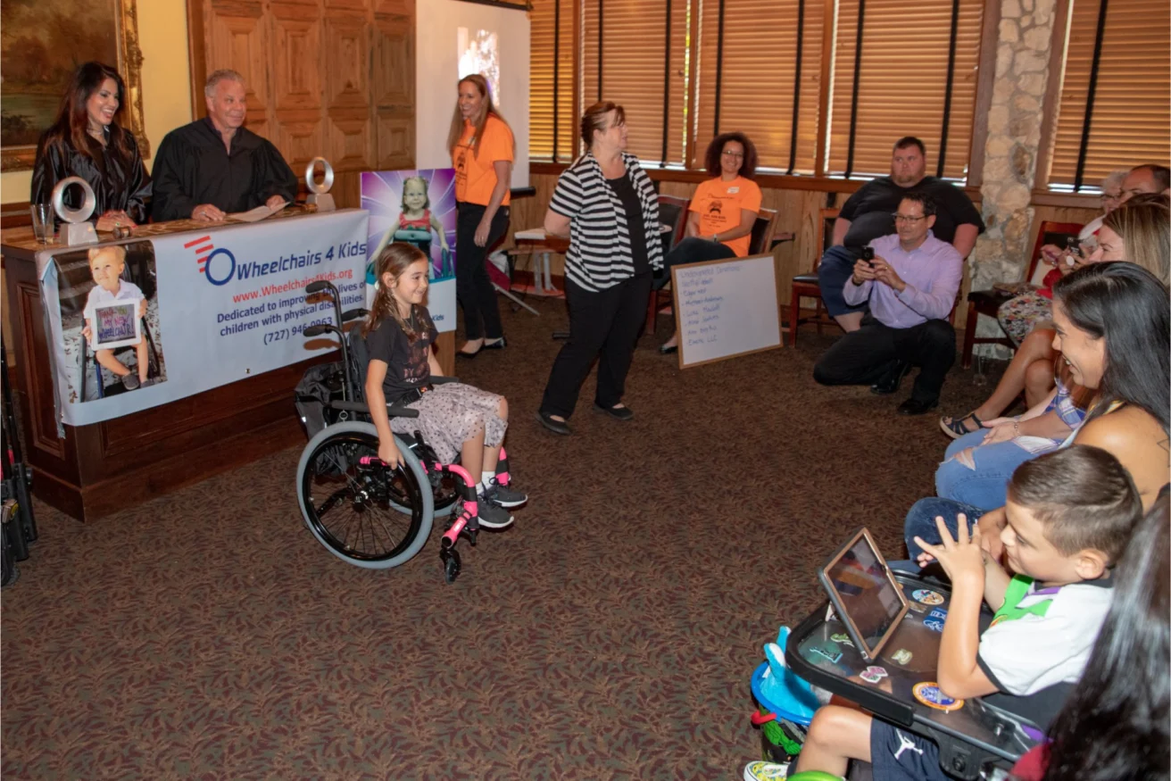 A girl in a wheelchair is at the center of a room with people around her. A banner reading "Wheelchairs 4 Kids" is displayed on the left and a woman is speaking at the front of the group.