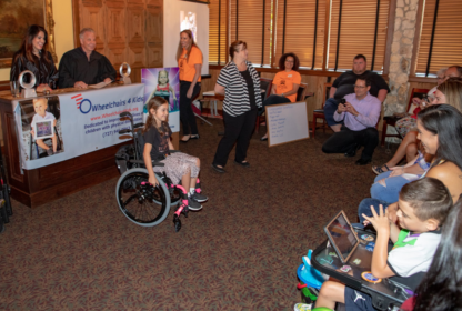A girl in a wheelchair is at the center of a room with people around her. A banner reading "Wheelchairs 4 Kids" is displayed on the left and a woman is speaking at the front of the group.