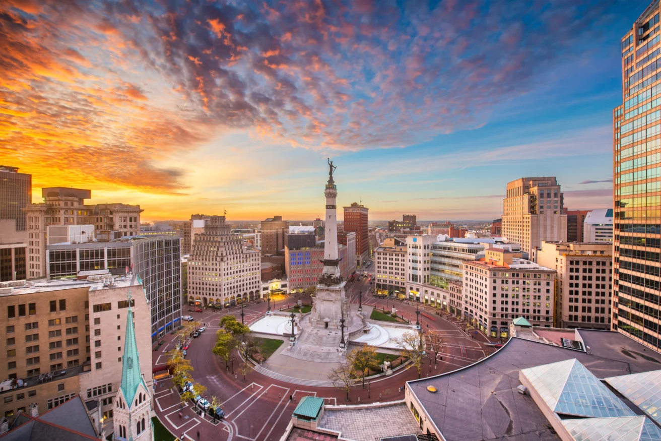 Aerial view of a city square at sunset with a central monument, several buildings surrounding the square, and a vibrant, colorful sky in the background.