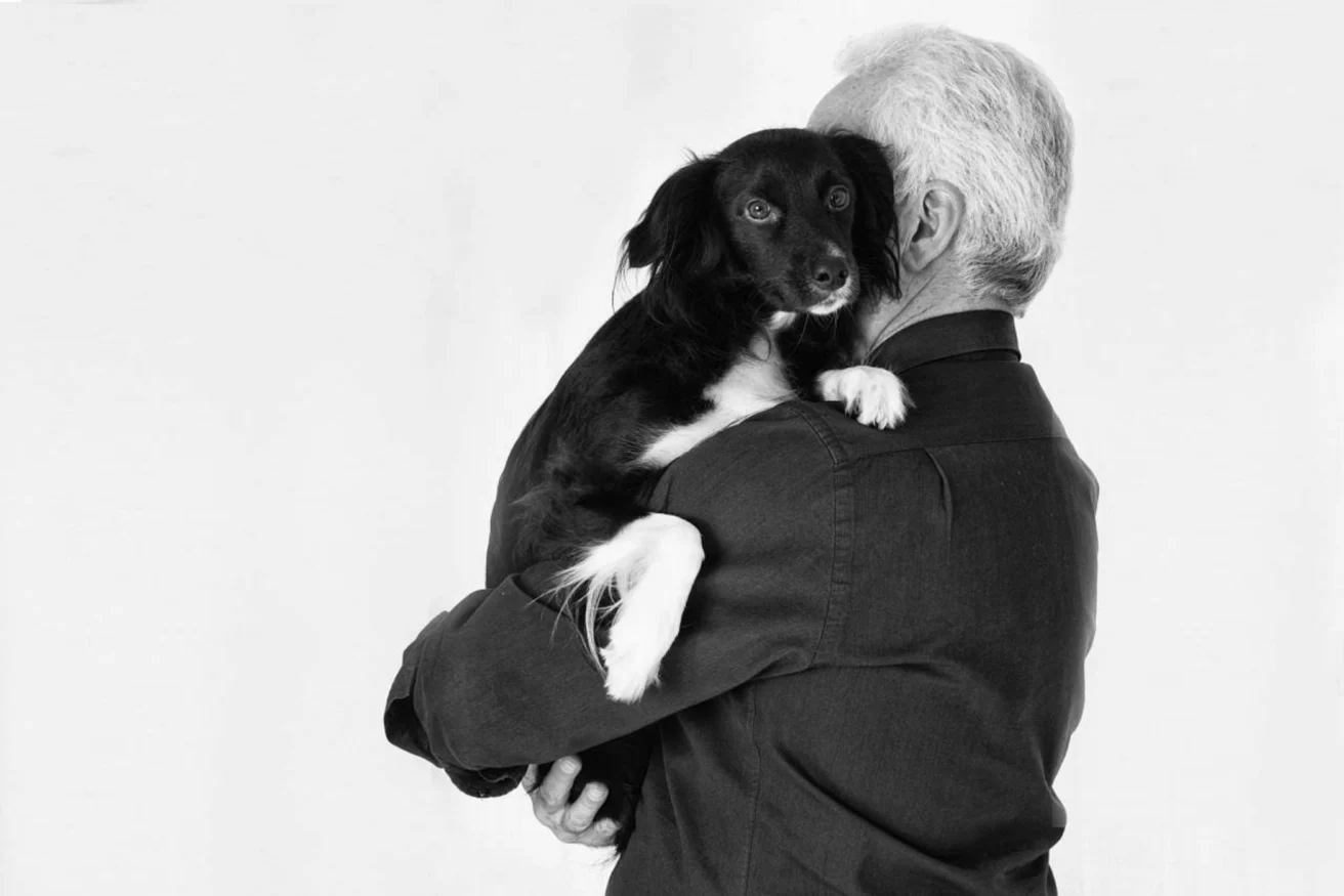 An elderly man with gray hair, seen from behind, holds a small black and white dog in his arms. The dog faces the camera. Image is in black and white.