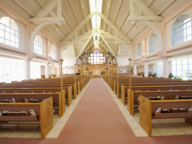 Interior of an empty church with wooden pews, a red carpet aisle, high vaulted ceiling, large windows, and a prominently displayed altar.