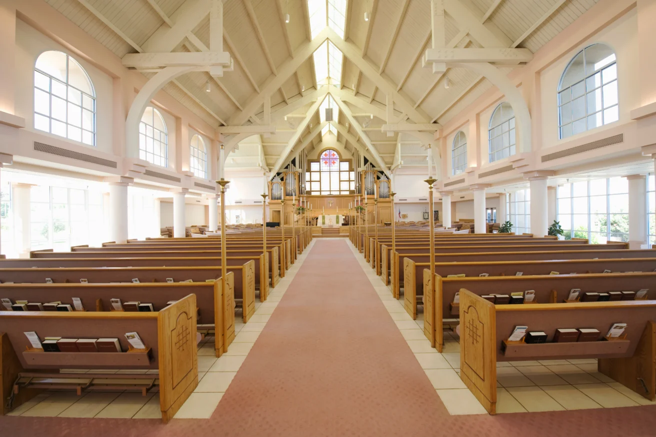 A bright, spacious church interior with wooden pews, high ceilings, large windows, and a central aisle leading to the altar.