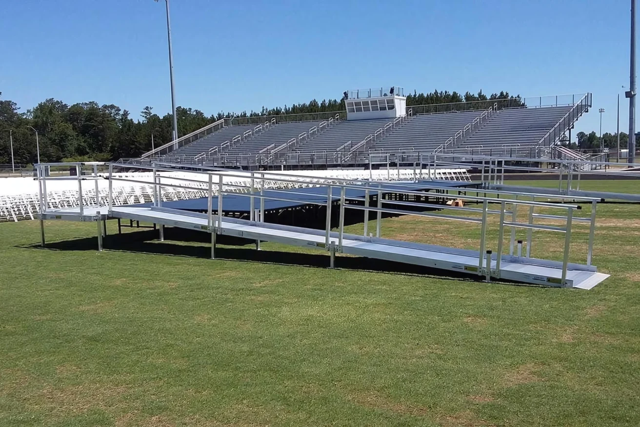 A large, empty stadium with metal bleachers and a metal ramp leading to the field, surrounded by green grass and clear skies.