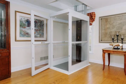A modern residential home elevator with glass doors is installed in a elegantly decorated room with paintings, a wooden floor, and a table.